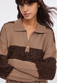 Polo in maglia con una striscia pelosa marrone e marrone scuro, caratterizzata da una chiusura con bottoni dorati e maniche testurizzate. Materiale morbido e accogliente.