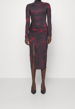 Robe à manches longues de couleur violet foncé avec un motif floral rouge, fronces sur le côté et fente frontale à la cuisse, portée avec des escarpins noirs à bouts pointus.