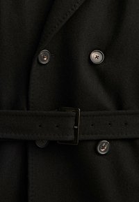 Gros plan d’un manteau en laine noir avec quatre boutons foncés et une ceinture assortie avec une boucle métallique rectangulaire.