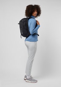 Zwarte rugzak met verstelbare schouderbanden, gedragen op de rug. Model in een lichtblauw langemouwen shirt en lichtgrijze broek, met grijze sneakers.