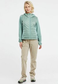 Šviesiai mėlyna izoliuota striukė su kapišonu, vertikaliu siuvinėjimu ir raštuotomis rankovėmis; derinama su bežinėmis kelnėmis ir daugiaspalviais batais.