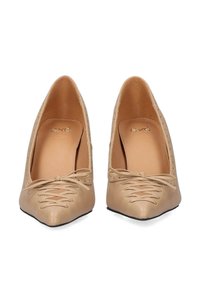 Zapatos de tacón beige de punta con un acabado de cuero suave, que presentan un detalle de cordones y pequeñas perforaciones a lo largo de los lados.