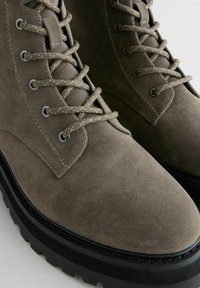 Braune Wildleder-Stiefeletten mit strukturierter Oberfläche, Metallösen und passendem Schnürsenkel. Dicke schwarze Gummisohle mit geriffeltem Profil.