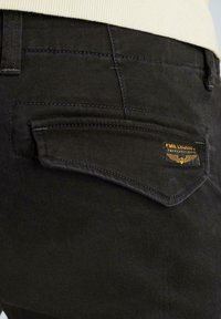 Schwarze Denim-Hose mit einer markanten schrägen Gesäßtasche, ausgestattet mit einem Lederetikett mit Text und Flügel-Icon. Glatte Textur und saubere Nähte.