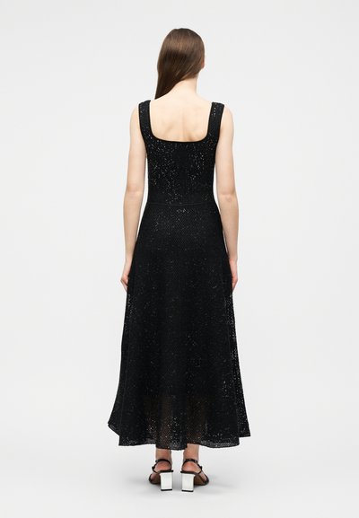 Γυναίκα με μακριά μαλλιά που φοράει μαύρο μακρύ φόρεμα με δίχτυ χωρίς μανίκια και μαύρα σανδάλια με λευκές χοντρές τακούνια, όρθια με την πλάτη προς το μέρος μας.