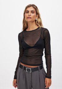 Svart genomskinlig långärmad topp med rund halsringning, med en underlagdesign. Kombination med högmidjade grå byxor och ett svart bälte.