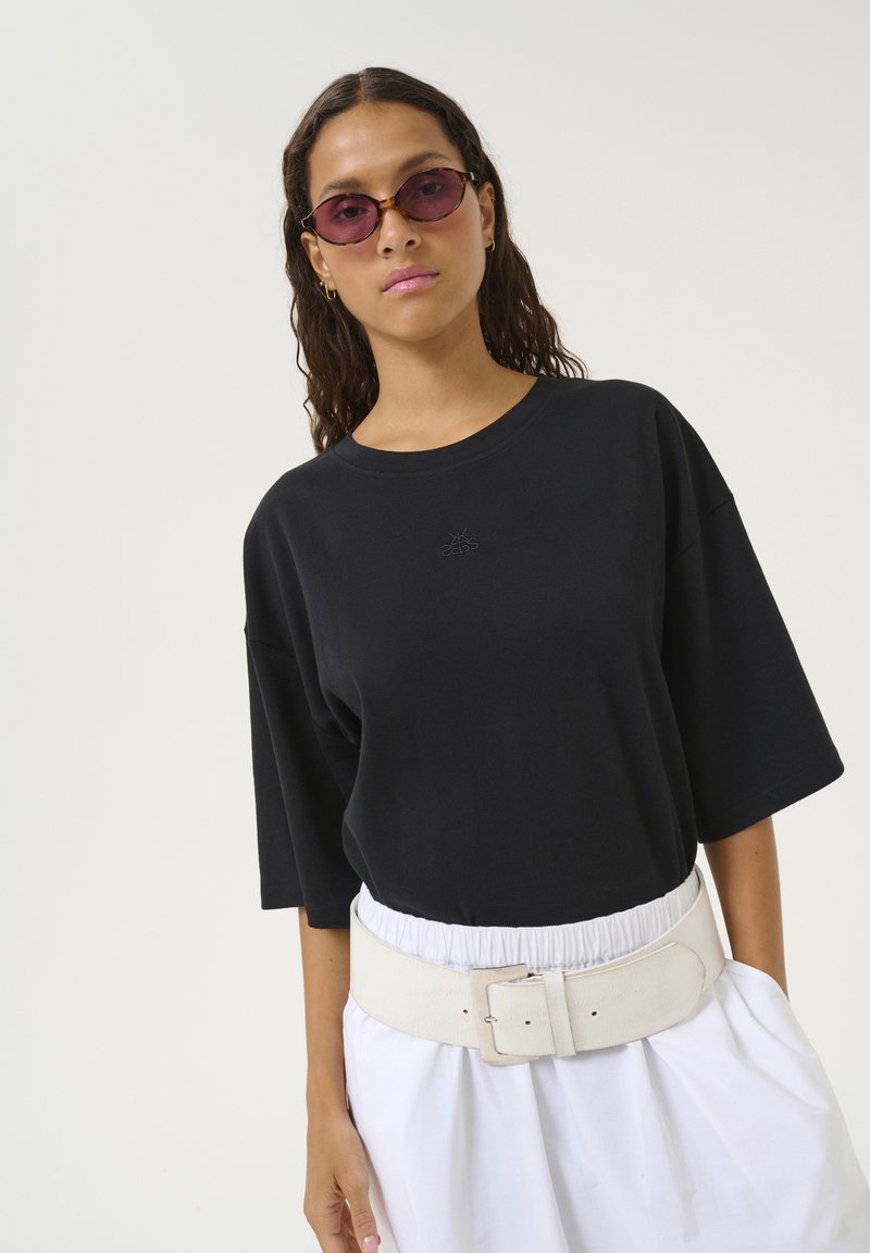 Sort oversized t-shirt med korte ærmer og subtile broderede logo, parret med et bredt hvidt bælte og løse hvide shorts.