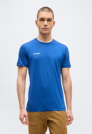 Junger Mann mit kurzen Haaren, der ein blaues Mammut-T-Shirt und beige Cargohosen trägt, steht vor einem schlichten hellen Hintergrund.