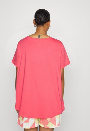 Person med hår sat op i en knold iført en løs lyserød T-shirt og shorts med geometrisk mønster, set bagfra mod en ensfarvet baggrund.