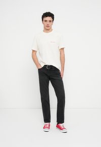 Εκρού μπλουζάκι με κόκκινο κείμενο, μαύρο τζιν παντελόνι και κόκκινα αθλητικά παπούτσια. Το μπλουζάκι έχει στρογγυλή λαιμόκοψη και το τζιν είναι στενό με ζώνη.