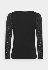 Chemise noire à manches longues avec détails en dentelle sur les manches et tissu uni sur le corps, sur un fond uni.