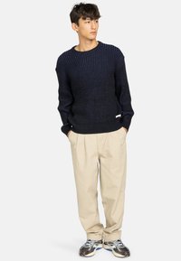 Maglione navy a coste con finitura testurizzata, scollatura rotonda e polsini aderenti, abbinato a pantaloni beige a pieghe e scarpe da corsa grigie.
