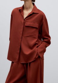 Rostfärgad skjorta med knappar, bröstficka, avslappnad passform, lätt tyg, långa ärmar och sidoflikar för större rörlighet.