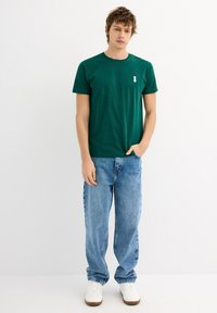 Camiseta de algodón verde con cuello redondo y un pequeño emblema blanco, combinada con unos vaqueros sueltos de color azul claro y zapatillas blancas.