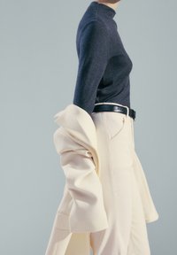 Personne portant un col roulé bleu foncé rentré dans un pantalon crème taille haute avec une ceinture noire, tenant un blazer crème drapé sur son bras.