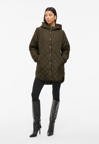 Manteau matelassé vert olive avec capuche, boutons pression et poches latérales, associé à des leggings noirs et des bottes pointues jusqu'aux genoux.