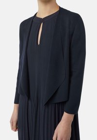 Granatowy dzianinowy bolerko z otwartym przodem i rękawami do połowy łokcia, zestawione z plisowaną ciemną sukienką. Miękka faktura, dopasowany krój, gładkie wykończenie.