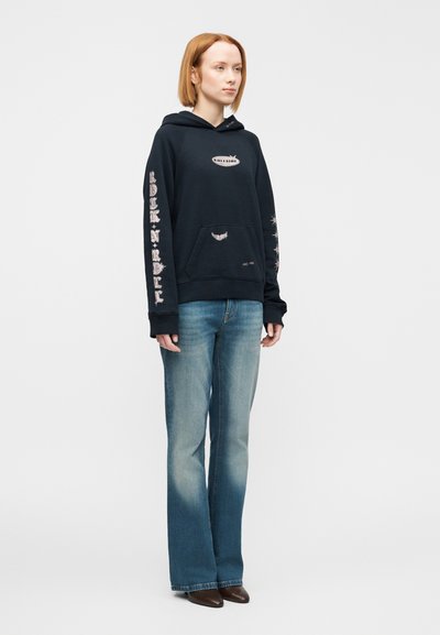 Μπλε σκούφος με τσέπη καγκουρό, λευκά τυπωμένα σχέδια στα μανίκια και στο στήθος, σε συνδυασμό με ανοιχτό μπλε καμπάνα τζιν και καφέ παπούτσια.