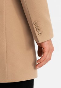 Blazer beige testurizzato con una vestibilità su misura, caratterizzato da un polsino a quattro bottoni e dettagli di cucitura pulita. Il tessuto presenta un sottile motivo a trama.