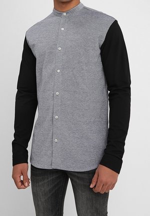 Man wearing a button-up shirt with a grey body and black long sleeves, paired with dark jeans, standing with hands relaxed.