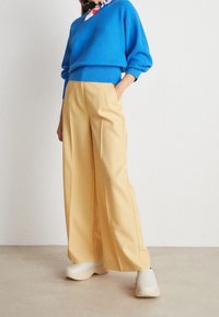 Femme portant un pull bleu texturé, un pantalon jaune à jambes larges, des chaussures blanches sans lacets et un foulard coloré autour du cou, debout contre un mur blanc uni.