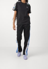 Svart bomullströja med blomsterbroderi och tre vita ränder på ärmarna, kombinerad med svarta byxor med blå och vita sidor.