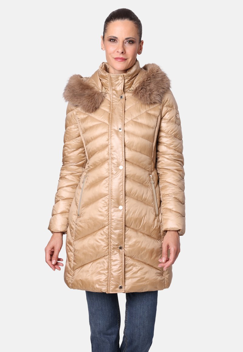 Mujer vestida con un abrigo de invierno acolchado beige con capucha de pelaje y bolsillos con cremallera, combinado con jeans azules, de pie contra un fondo blanco.