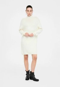 Rochie tricotată de culoare crem, cu guler înalt și mâneci lungi, asortată cu botine negre cu accente de catarame.