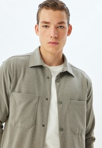 Ljusgrå button-up skjorta i texturerad väv, med två framfickor, krage och en enkel vit undertröja.