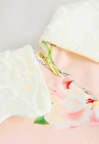 Pastelovo ružová tkanina s kvetinovým vzorom, biely textúrovaný golier a zlatý zips. Obsahuje jemnú, hladkú textúru a delikátne dizajnové prvky.