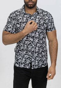 Chemise noire à manches courtes avec un motif floral blanc. Design à boutons avec un col et un ourlet droit. Portée sur un pantalon noir.