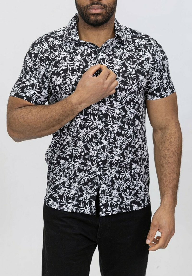 Chemise noire à manches courtes avec un motif floral blanc. Design à boutons avec un col et un ourlet droit. Portée sur un pantalon noir.
