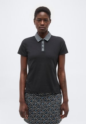 Žena oblečená v čiernej polo košeli s krátkym rukávom a sivým vzorovaným golierom, a v čiernej sukni s bielym a modrým abstraktným vzorom.