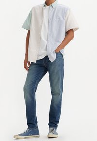 Man som bär en kortärmad, knappad, flerfärgad randig skjorta över en vit t-shirt, blå jeans och blåa högsneakers.