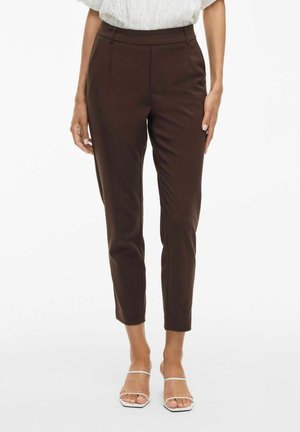 Brązowe spodni z wysokim stanem, slim fit, minimalistyczne detale w postaci zagnieceń i długości do kostek, zestawione z białymi sandałami na paski.