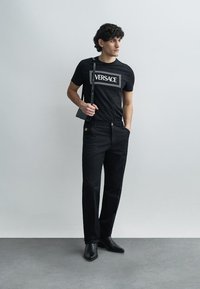 T-shirt preta de algodão com o logotipo branco "VERSACE", combinada com calças pretas que têm um botão dourado e um bolso lateral. Sapatos pretos completam o visual.