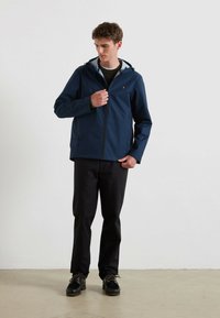 Granatowa kurtka przeciwdeszczowa z zamkiem z przodu, regulowanym kapturem, bocznymi kieszeniami oraz z logo na piersi, w połączeniu z czarnymi spodniami i butami.