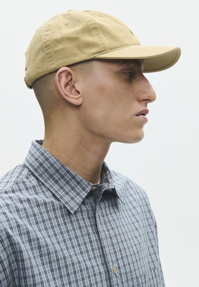 Mies beige baseball-lipulla ja siniharmaa ruutupaita katsoo oikealle yksiväristä valkoista taustaa vasten.