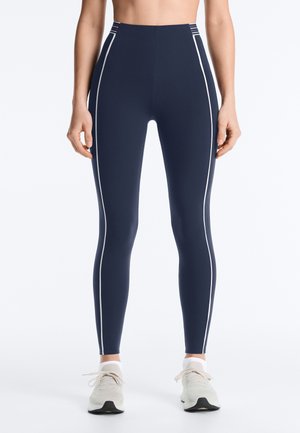 Person trägt marineblaue hoch taillierte Leggings mit weißen Seitenstreifen und weiße Turnschuhe, steht vor einem schlichten Hintergrund.