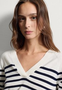 Maglione a righe in crema con righe orizzontali blu navy, con scollatura a V e colletto a coste. Texture morbida e design aderente.