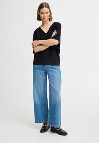 Haut noir à manches courtes et décolleté en V associé à un jean bleu à jambes larges. La tenue présente une coupe décontractée et un design minimaliste. Des mocassins noirs complètent le look.