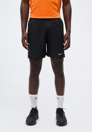 Hombre que lleva puestos unos pantalones cortos negros de Nike, calcetines blancos Nike hasta la mitad de la pierna, zapatillas negras y una camiseta naranja, de pie con los brazos relajados a los lados.