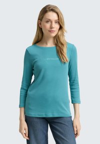 Türkise Langarmshirt mit rundem Ausschnitt und einfachem Textlogo in Weiß. Aus glattem Baumwollstoff, figurbetonter Silhouette, kombiniert mit Jeans.