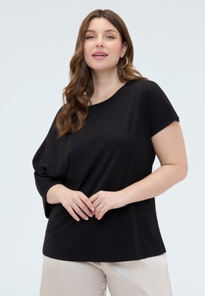 Kvinde med langt brunt hår iført en sort asymmetrisk top med korte ærmer og lyse bukser, stående med hænderne foldet.