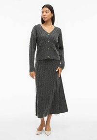 Cardigan grigio a maglia con scollo a V e cinque bottoni, abbinato a una gonna lunga coordinata, entrambi strutturati, con motivi a righe verticali.