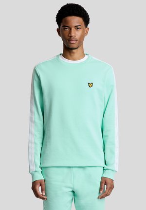 Jonge man draagt een licht turquoise sweatshirt en bijpassende broek met een geel vogellogo op de borst, staande tegen een grijze achtergrond.
