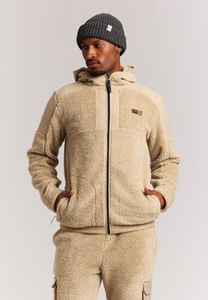 Man met een beige fleecejack en broek met handen in de zakken en een donkergrijze gebreide muts, kijkt naar links.