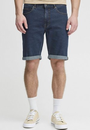 Denim kratke hlače v temno modri barvi z zavihanimi robovi, ki imajo pet žepov in klasičen dizajn z zadrgo, stilizirane s bež športnimi copati in belimi nogavicami.