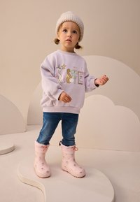 Lavandas krāsas džemperis ar krāsainu grafisku dizainu, zili džinsi un rozā ūdensizturīgi zābaki ar ziedu rakstu, nēsāti kopā ar gaišu adītu cepuri.