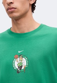 Άνδρας που φοράει πράσινο μπλουζάκι των Boston Celtics με μασκότ να περιστρέφει το μπάσκετ στον δείκτη του και το λογότυπο της Nike από πάνω.